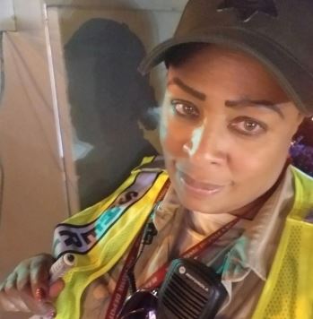 Deputy Harris is looking at the camera selfie style. Dressed in Sheriff uniform tan shirt with a radio off her left collar. wearing a yellow safety vest.